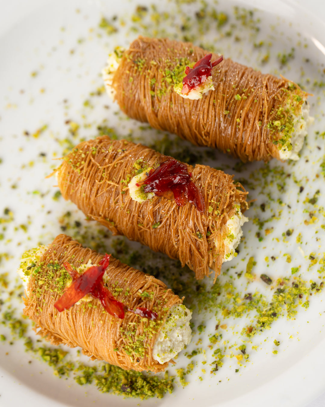 Three Esmaliye rolls filled with sweet ashta, garnished with pistachios and rose petal on white plate