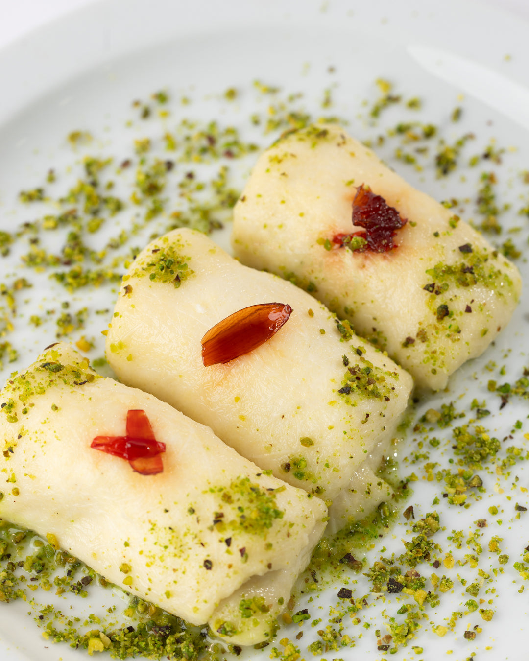 Three creamy Halewet el Jeben rolls garnished with ground pistachios and flower jam on a white plate.