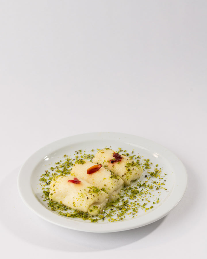 Three creamy Halewit el Jeben rolls garnished with ground pistachios and floral jam on a white plate.